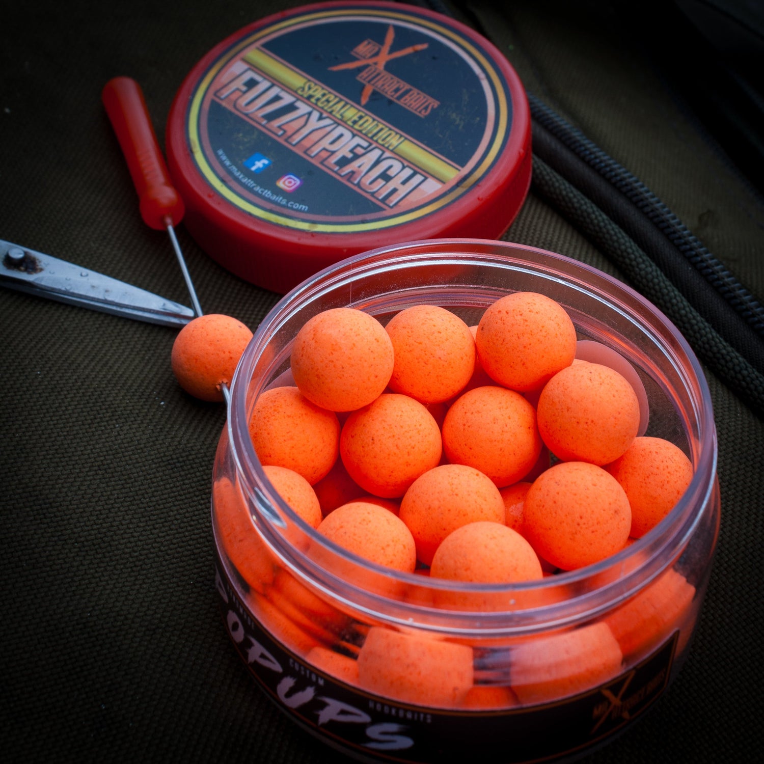 pot of orange max attract baits fuzzy peach popups on a bag with the lid behind, a bating needle next to the pot with a popup on the needle
