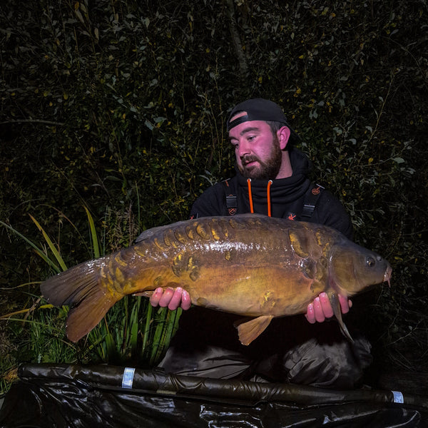 angler crouched down holding large mirror carp over a mat at night with green reeds and trees behind him wearing max attract baits hoodie and cap