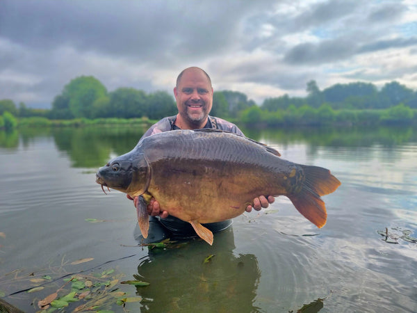 angler stood in carp lake holding a big mirror carp with a green tree line behind him