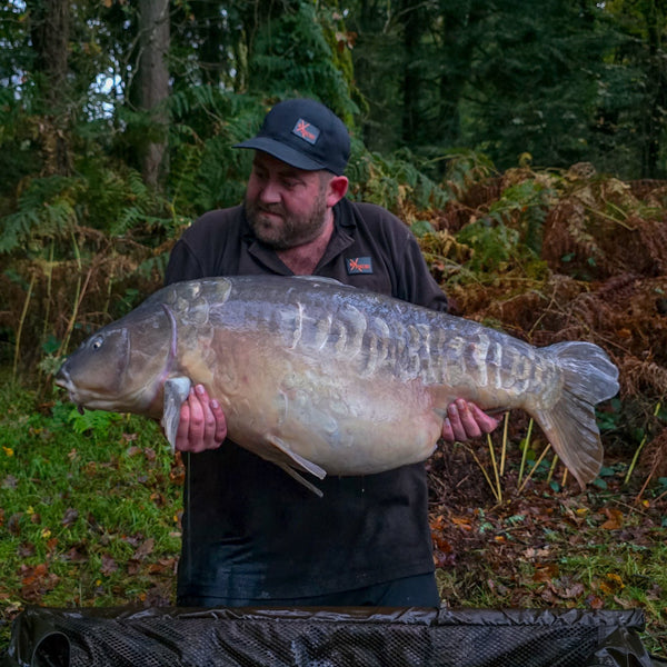 angler holding large mirror carp over a mat with a wooded background behind wearing max attract baits tshirt and cap