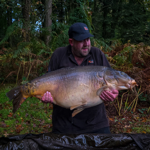 angler kneeling holding big carp set in a wooded background wearing max attract baits tshirt and cap
