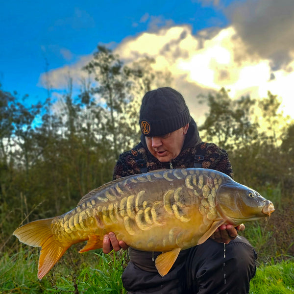 angler holding mirror carp with grass and trees behind him wearing max attract baits fleece and bobble hat