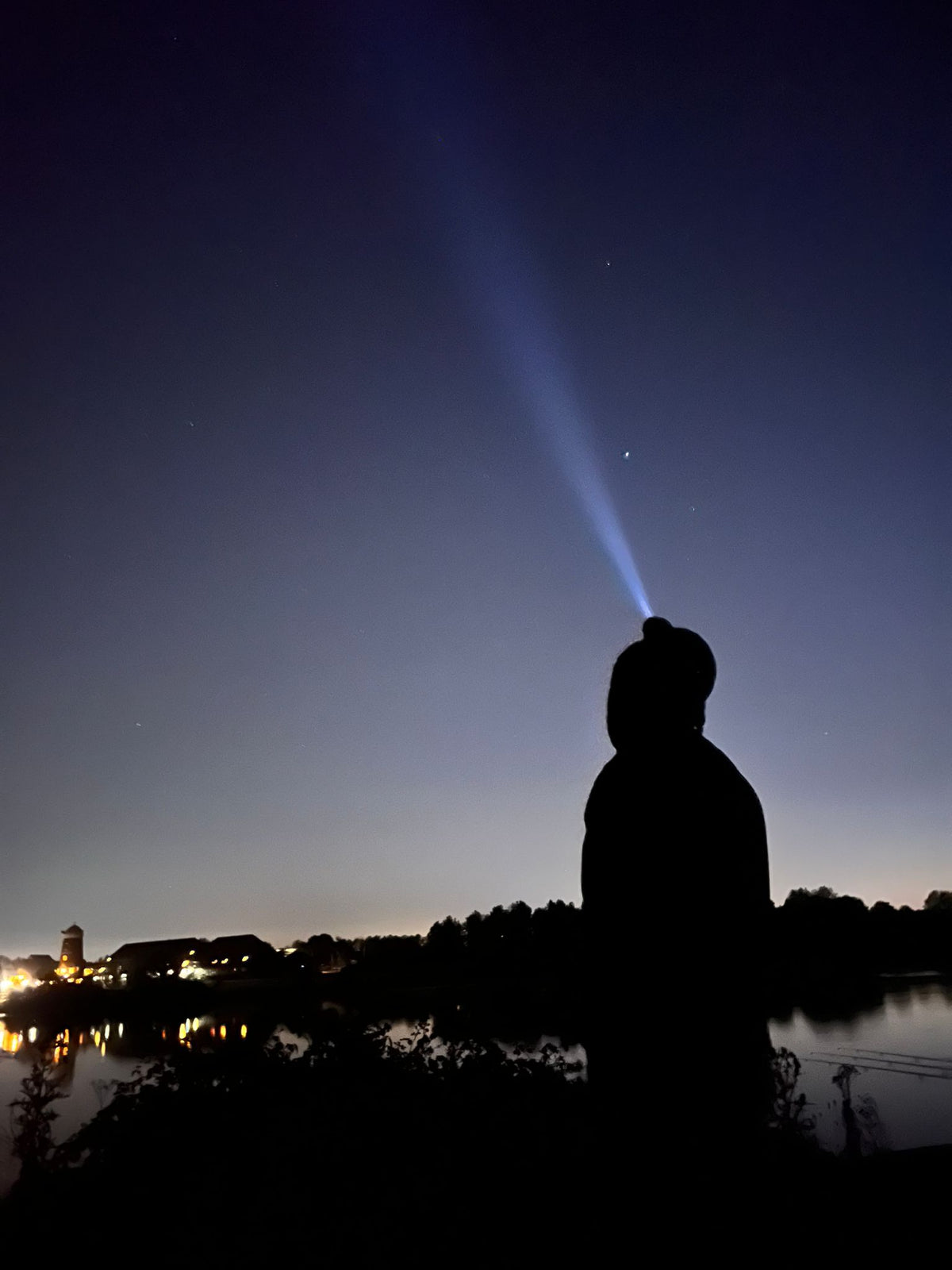 angler with head torch on head looking up at the night sky in a silloutte with town lighs in back ground. 