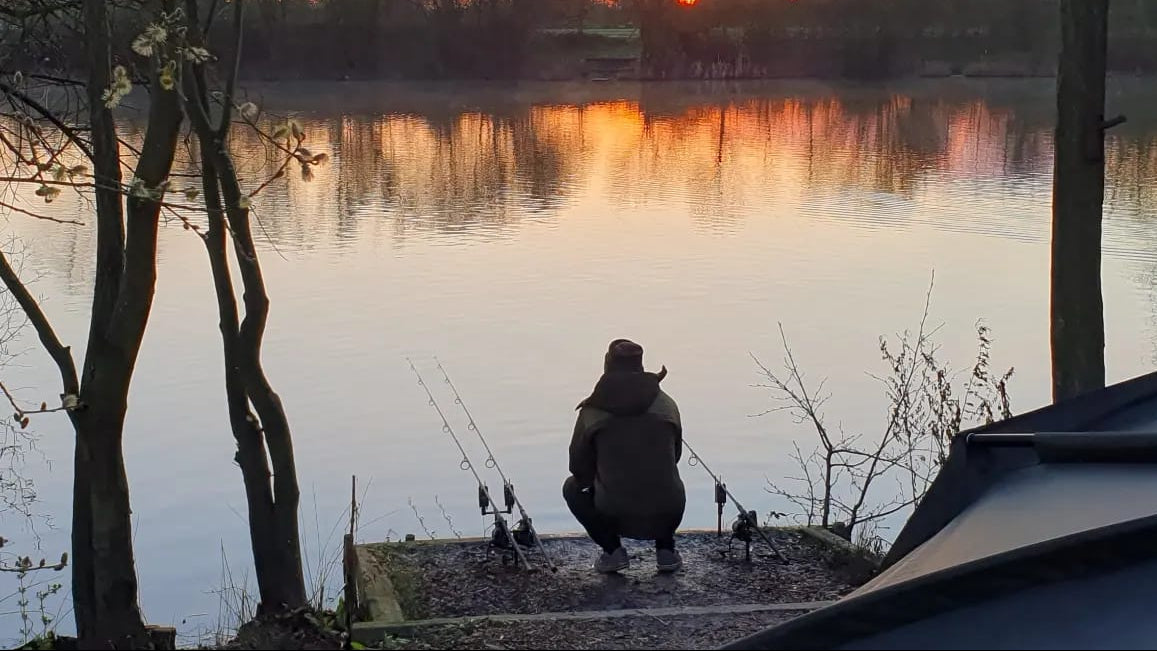 Angler knelt next to rods watching the lake as the sun sets.
