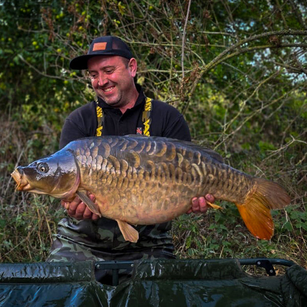 angler cradling a large mirror carp set in a wooded background with slight greenery wearing max attract baits cap