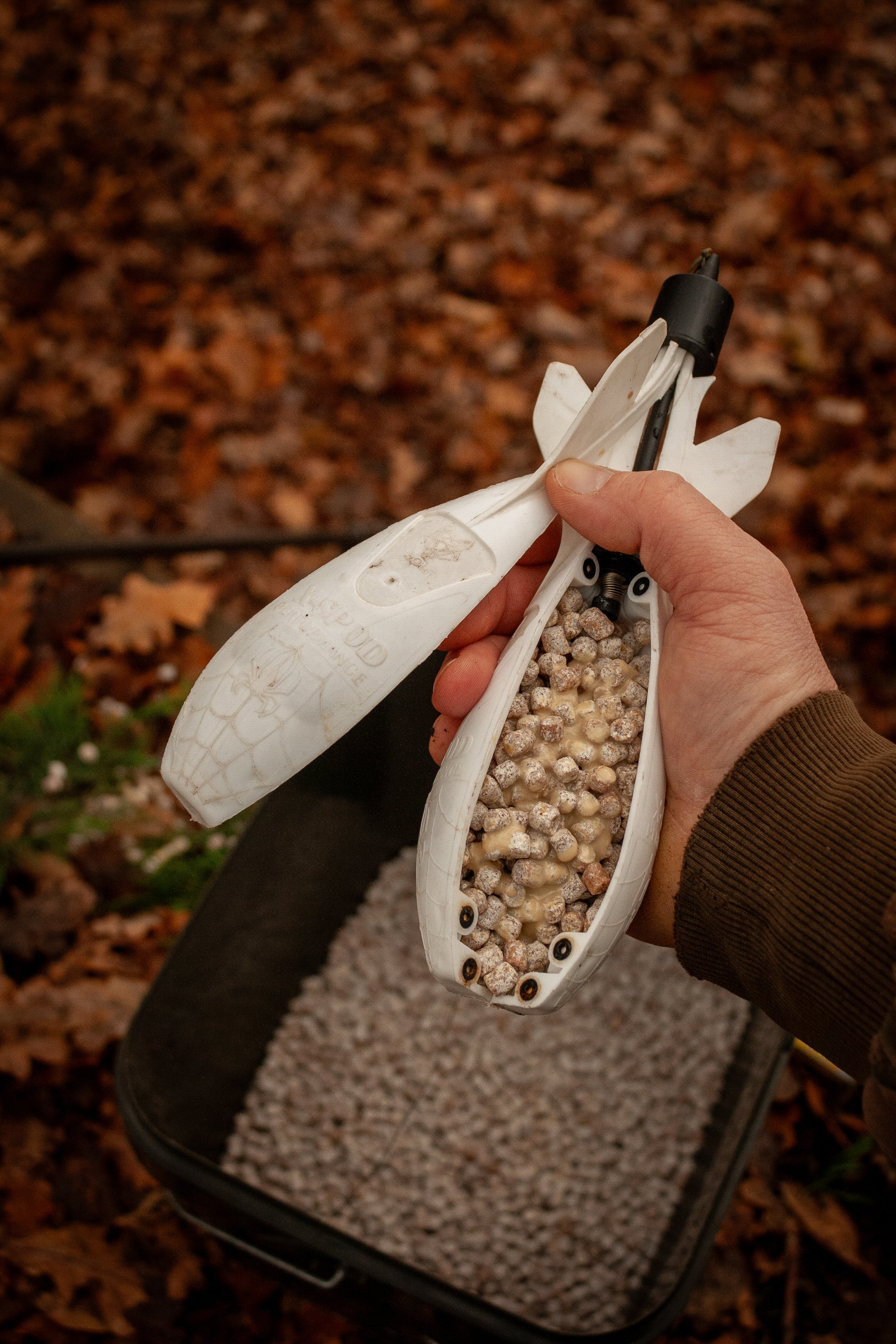person holding a spomb full of max attract baits pinacolada pellets with a bucket of pellets underneath set in a leafy autumnal back ground