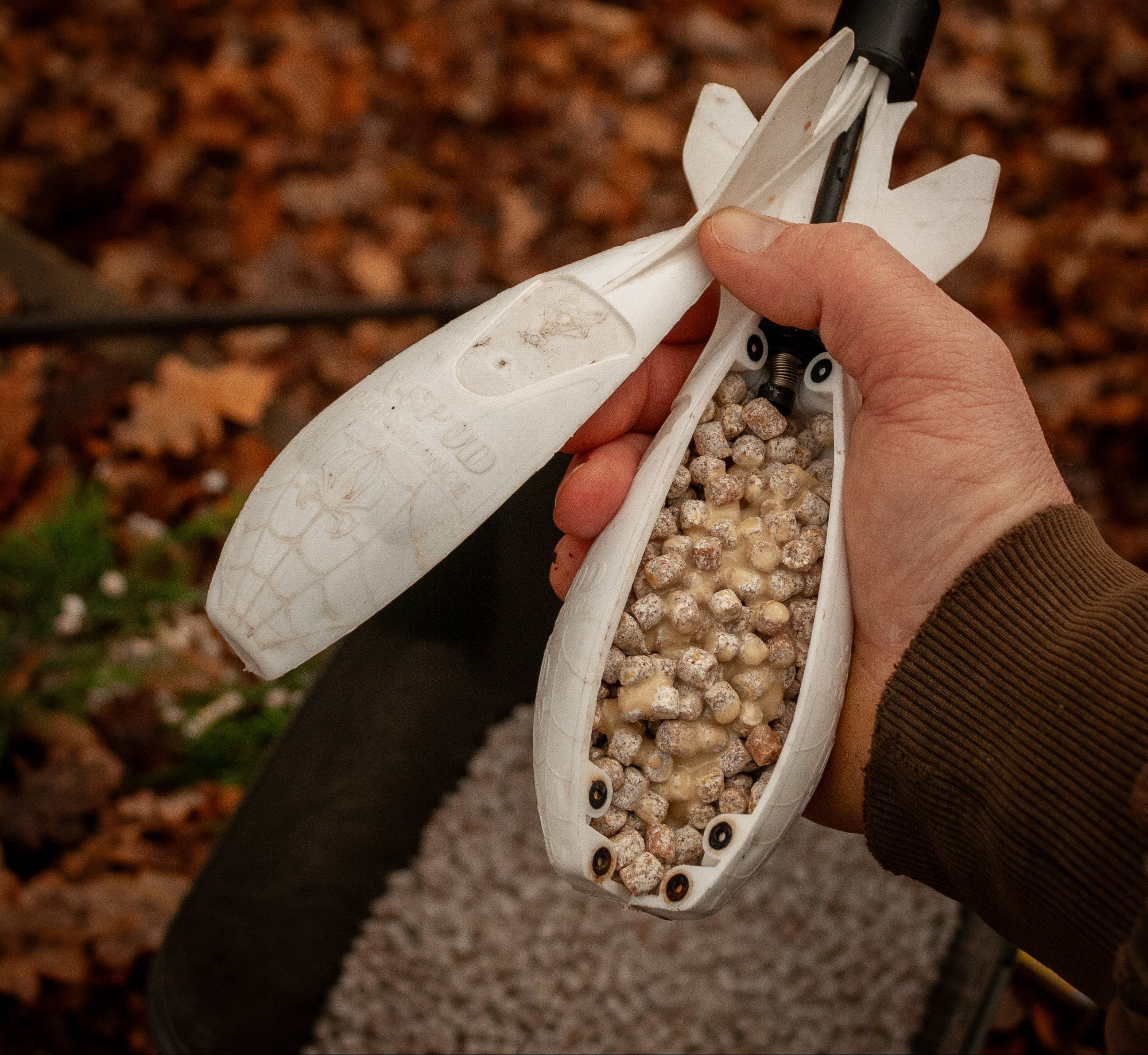 person holding a spomb full of max attract baits pinacolada pellets with a bucket of pellets underneath set in a leafy autumnal back ground